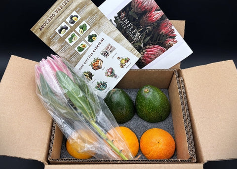 Open cardboard box containing avocados, oranges, a bouquet of flowers, and a card with plant illustrations.
